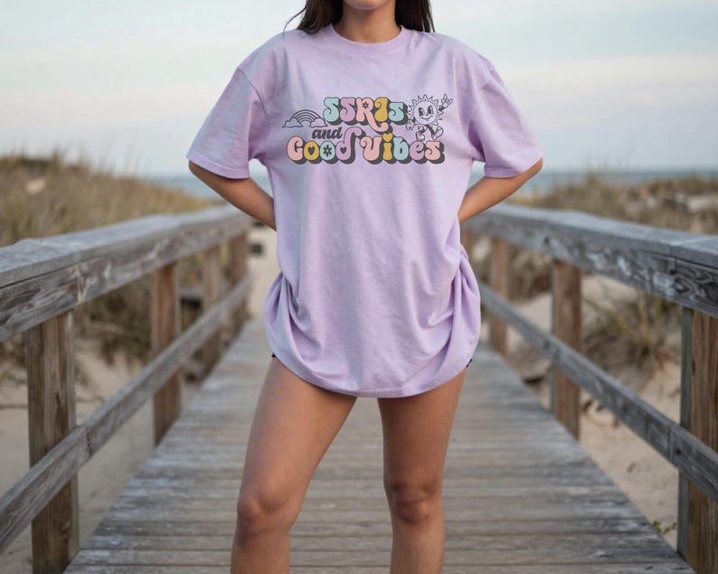 May include: A light purple t-shirt with the text "SSR's and Good Vibes" in a retro font. The graphic includes a rainbow, sun, and other elements. The shirt is a relaxed fit.