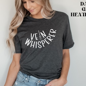 以下が含まれることがあります： 暗いグレーのヘザーTシャツに、白い文字で「Vein Whisperer」と書いてあります。テキストは半円形に曲がっており、中央には小さな針と注射器のグラフィックがあります。