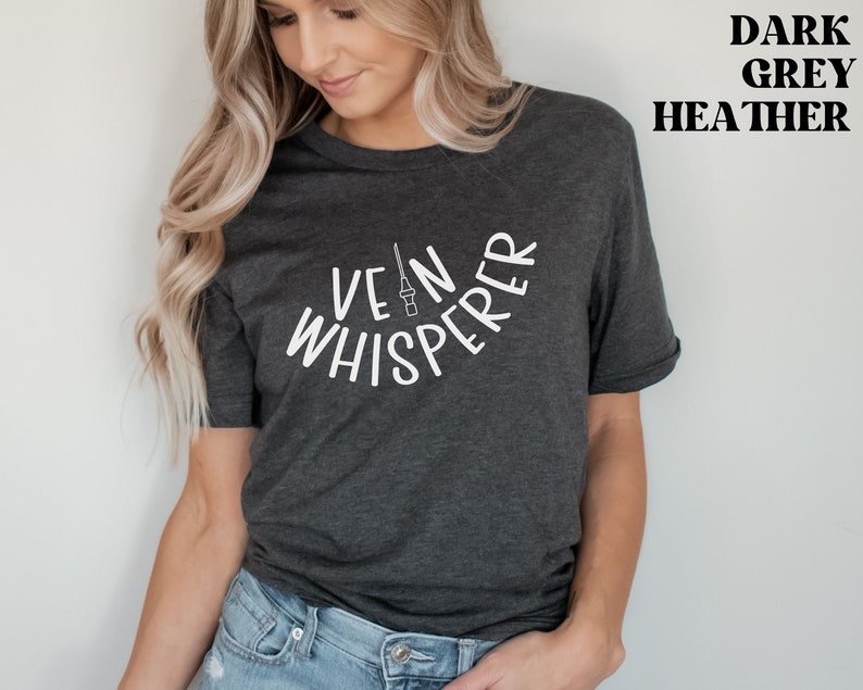 以下が含まれることがあります： 暗いグレーのヘザーTシャツに、白い文字で「Vein Whisperer」と書いてあります。テキストは半円形に曲がっており、中央には小さな針と注射器のグラフィックがあります。