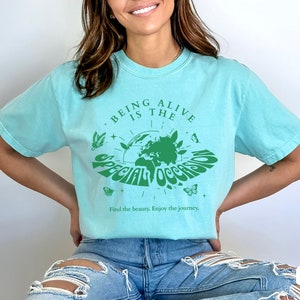 May include: A turquoise blue t-shirt with a green graphic design featuring a globe with the text "Being Alive is the Special Occasion" and the words "Find the beauty. Enjoy the journey."