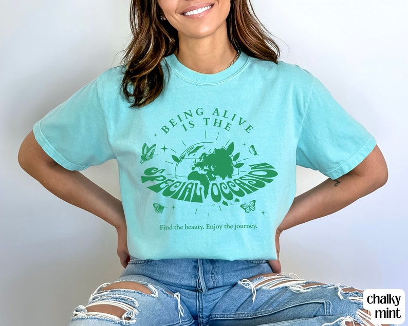 May include: A turquoise blue t-shirt with a green graphic design featuring a globe with the text "Being Alive is the Special Occasion" and the words "Find the beauty. Enjoy the journey."