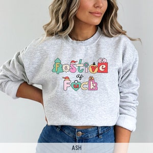 Op de afbeelding: Een grijs sweatshirt met een feestelijk ontwerp dat "Festive as f*ck" zegt in kleurrijke letters en illustraties. Het ontwerp bevat een kerstboom, een cadeaudoos, een kerstmanmuts en een hart.