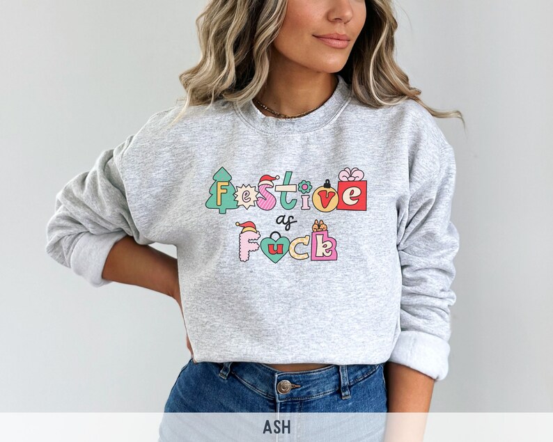 Op de afbeelding: Een grijs sweatshirt met een feestelijk ontwerp dat "Festive as f*ck" zegt in kleurrijke letters en illustraties. Het ontwerp bevat een kerstboom, een cadeaudoos, een kerstmanmuts en een hart.