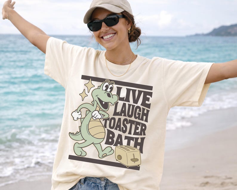 May include: Cream-colored t-shirt featuring a cartoon alligator and toaster with the words "LIVE LAUGH TOASTER BATH" in black. The alligator is light green with a beige belly and is walking. The toaster is beige.