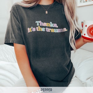 May include: A dark gray t-shirt with the text "Thanks, it's the trauma." in colorful letters.