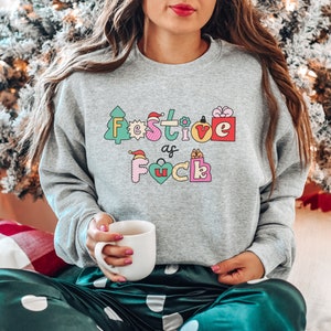 Op de afbeelding: Een grijs sweatshirt met een feestelijk ontwerp dat "Festive as f*ck" zegt in kleurrijke letters met kerstthema-illustraties.