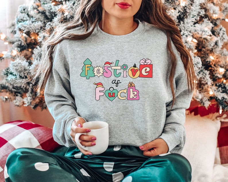 Op de afbeelding: Een grijs sweatshirt met een feestelijk ontwerp dat "Festive as f*ck" zegt in kleurrijke letters met kerstthema-illustraties.