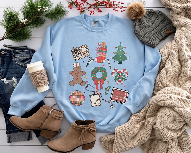 May include: Light blue sweatshirt with a Christmas-themed design featuring a gingerbread person, a stocking, a Christmas tree, a wreath, candy canes, a stethoscope, a bandage, a disco ball, a medical chart, and an EKG monitor.