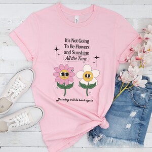 May include: Pink t-shirt with a graphic of two cartoon flowers and the text "It's Not Going To Be Flowers and Sunshine All the Time But they will be back again".