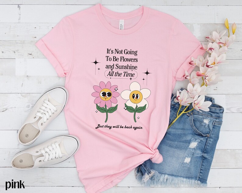 May include: Pink t-shirt with a graphic of two cartoon flowers and the text "It's Not Going To Be Flowers and Sunshine All the Time But they will be back again".