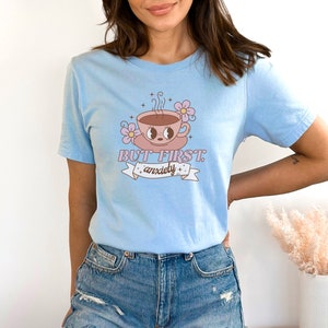 May include: Light blue t-shirt with a cartoon illustration of a coffee cup with eyes and a pink flower. The text "BUT FIRST, anxiety" is printed on the shirt.