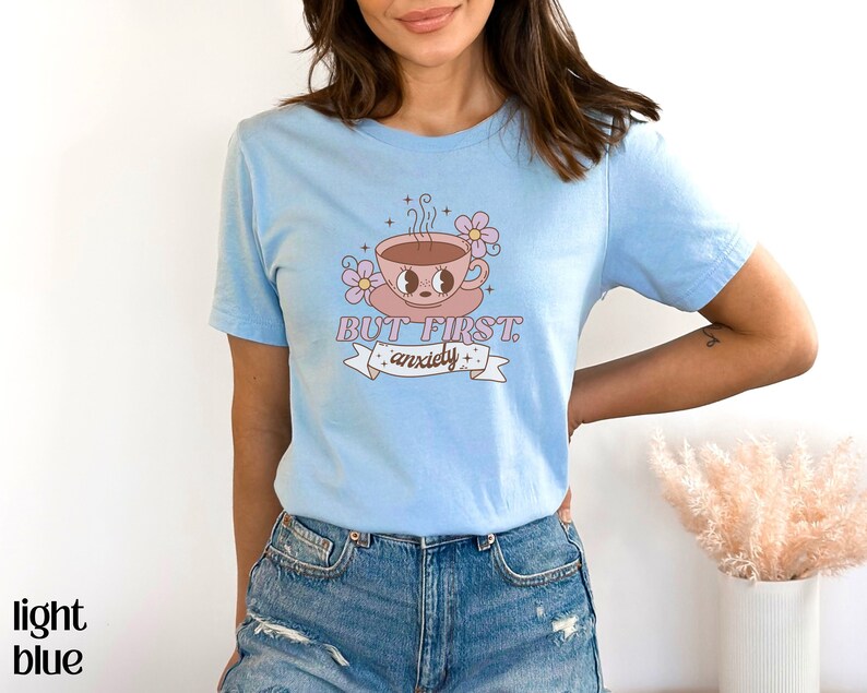 May include: Light blue t-shirt with a cartoon illustration of a coffee cup with eyes and a pink flower. The text "BUT FIRST, anxiety" is printed on the shirt.
