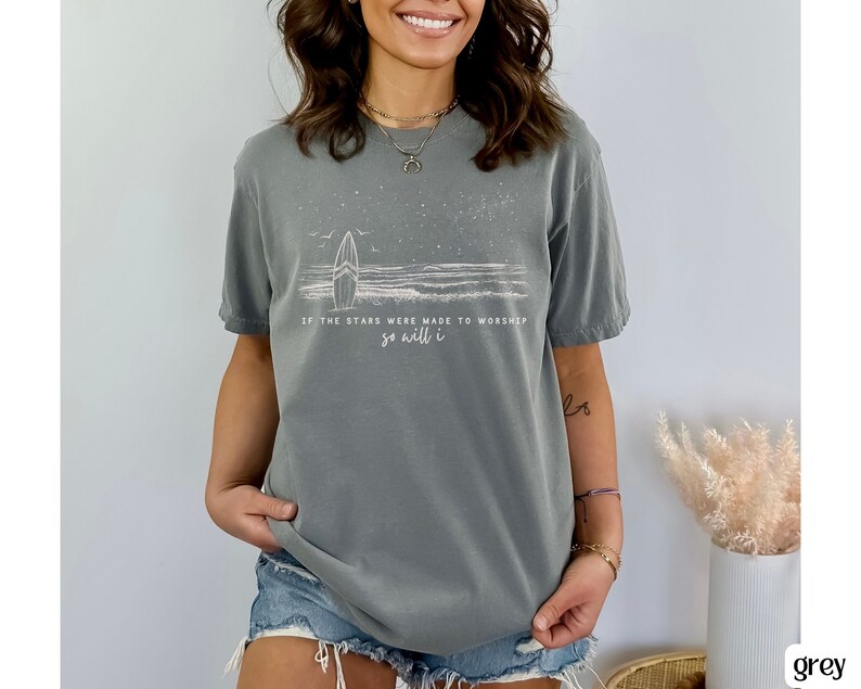 Puede incluir: Camiseta gris con un gr&aacute;fico blanco de una tabla de surf y olas con el texto "Si las estrellas fueron hechas para adorar, yo tambi&eacute;n lo har&eacute;".