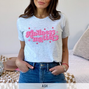 Pode incluir: Camiseta cinza claro com a frase "Kindness matters" em rosa, com letras retr&ocirc;. A camiseta tem gola redonda e mangas curtas. A modelo est&aacute; usando jeans azuis.