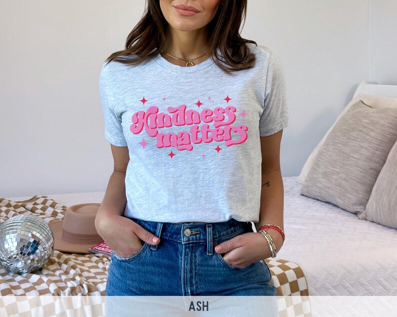 Pode incluir: Camiseta cinza claro com a frase "Kindness matters" em rosa, com letras retr&ocirc;. A camiseta tem gola redonda e mangas curtas. A modelo est&aacute; usando jeans azuis.