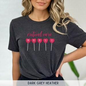 Pu&ograve; includere: Maglietta in jersey grigio scuro con il testo "critical care" e la parola "NURSE" scritta con lecca-lecca a forma di cuore rosa.