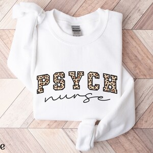 May include: White sweatshirt with the text "PSYCH nurse" in black script. The word "PSYCH" is in a block letter style with a leopard print pattern. The sweatshirt is folded on a light brown wooden surface.