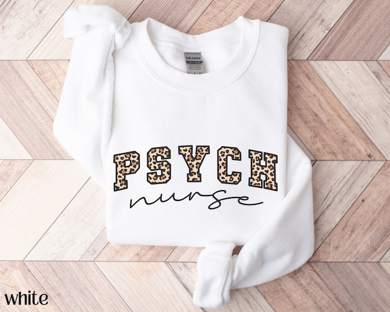 May include: White sweatshirt with the text "PSYCH nurse" in black script. The word "PSYCH" is in a block letter style with a leopard print pattern. The sweatshirt is folded on a light brown wooden surface.