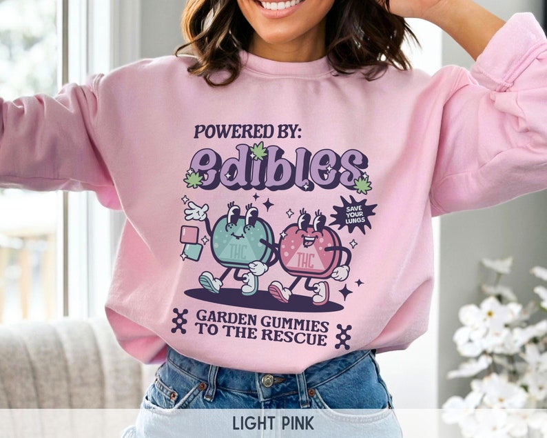 May include: A light pink sweatshirt with a graphic design featuring two cartoon gummies with the text "POWERED BY: edibles" and "GARDEN GUMMIES TO THE RESCUE".