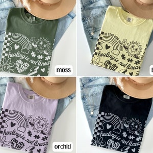 May include: Four t-shirts in different colours with a graphic print of a rainbow, a sun, a heart, a flower, a puzzle piece, and the text "Be your own sunshine", "healing is not linear", and "Find joy in the journey". The t-shirts are laid out on a denim background with a brown hat.