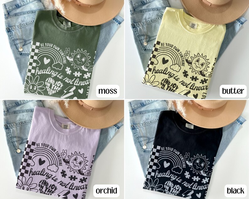 May include: Four t-shirts in different colours with a graphic print of a rainbow, a sun, a heart, a flower, a puzzle piece, and the text "Be your own sunshine", "healing is not linear", and "Find joy in the journey". The t-shirts are laid out on a denim background with a brown hat.