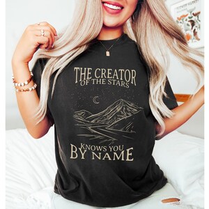 May include: Black t-shirt featuring a graphic of a mountain range, a crescent moon, and stars. The text "The Creator of the Stars Knows You By Name" is printed on the shirt.