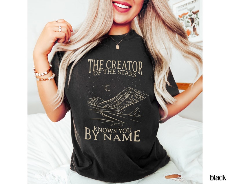 May include: Black t-shirt featuring a graphic of a mountain range, a crescent moon, and stars. The text "The Creator of the Stars Knows You By Name" is printed on the shirt.
