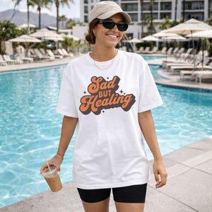 Puede incluir: Camiseta blanca con el texto "Sad but Healing" en fuente retro naranja y marr&oacute;n. La camiseta se usa con pantalones cortos negros y gafas de sol. El fondo incluye una piscina y palmeras.