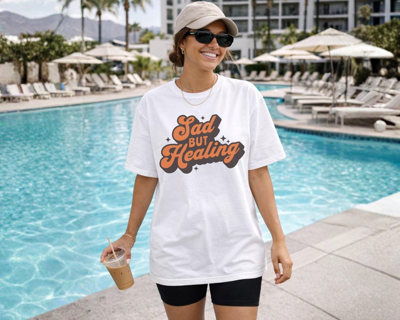 Puede incluir: Camiseta blanca con el texto "Sad but Healing" en fuente retro naranja y marr&oacute;n. La camiseta se usa con pantalones cortos negros y gafas de sol. El fondo incluye una piscina y palmeras.