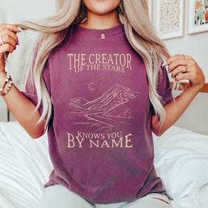 May include: A purple t-shirt with a graphic design of a mountain range, a crescent moon, and stars. The text "The Creator of the Stars Knows You By Name" is printed on the shirt.
