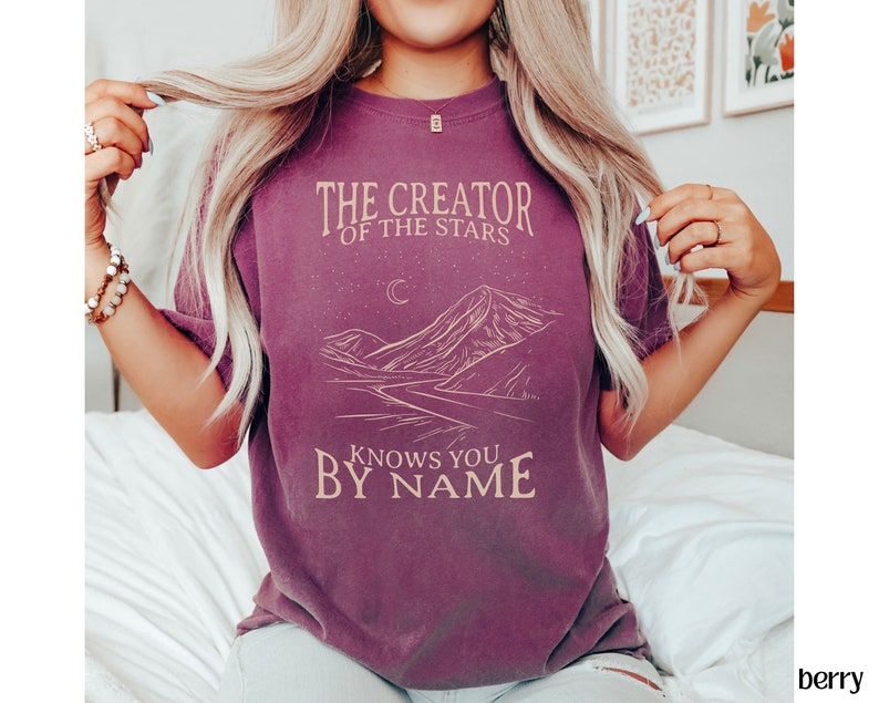 May include: A purple t-shirt with a graphic design of a mountain range, a crescent moon, and stars. The text "The Creator of the Stars Knows You By Name" is printed on the shirt.
