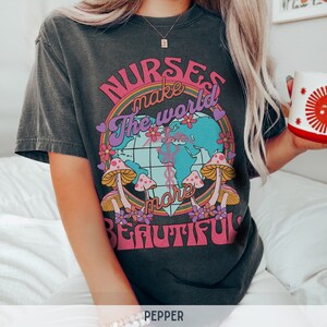 Puede incluir: Una camiseta gris oscuro con un dise&ntilde;o gr&aacute;fico colorido que presenta un globo terr&aacute;queo, hongos y flores. El texto en la camiseta dice "Nurses make the world more beautiful."