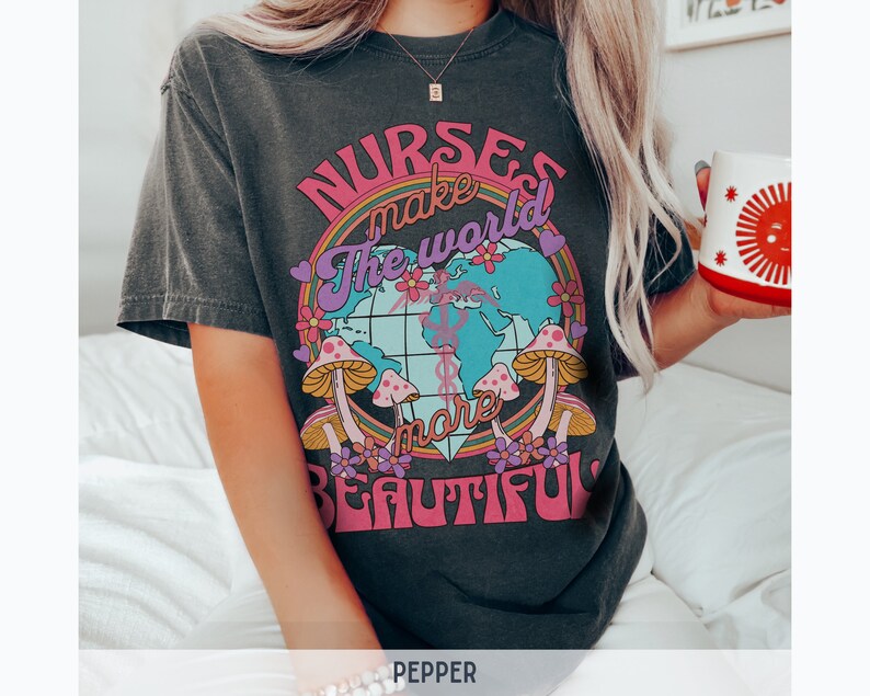 Puede incluir: Una camiseta gris oscuro con un dise&ntilde;o gr&aacute;fico colorido que presenta un globo terr&aacute;queo, hongos y flores. El texto en la camiseta dice "Nurses make the world more beautiful."