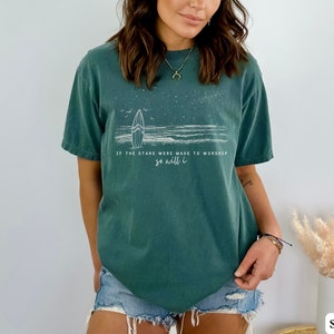 Puede incluir: Una camiseta azul turquesa con un gr&aacute;fico blanco de una tabla de surf y olas. El texto en la camiseta dice "Si las estrellas fueron hechas para adorar, yo tambi&eacute;n lo har&eacute;."