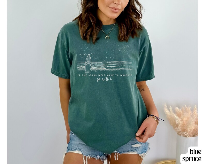 Puede incluir: Una camiseta azul turquesa con un gr&aacute;fico blanco de una tabla de surf y olas. El texto en la camiseta dice "Si las estrellas fueron hechas para adorar, yo tambi&eacute;n lo har&eacute;."