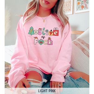 Op de afbeelding: Een lichtroze sweatshirt met de tekst "Festive as F*ck" in kleurrijke letters en kerstthema-afbeeldingen.