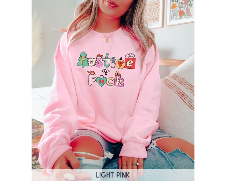 Op de afbeelding: Een lichtroze sweatshirt met de tekst "Festive as F*ck" in kleurrijke letters en kerstthema-afbeeldingen.