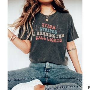 May include: A dark gray t-shirt with a vintage-style graphic that reads "Stars & Stripes & Running for Grill Lights". The graphic is in red, blue, and white with a firework design.
