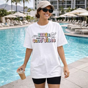 May include: White t-shirt with the text "SSRIs and Good Vibes" in colorful retro font. The shirt features a rainbow and a smiling sun graphic. The person is wearing black shorts, sunglasses, and a cap, holding a beverage near a pool.