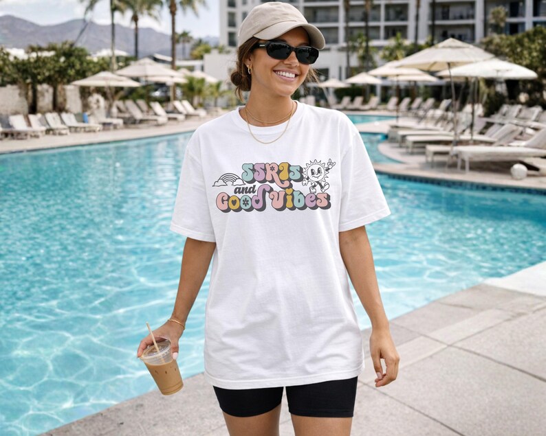 May include: White t-shirt with the text "SSRIs and Good Vibes" in colorful retro font. The shirt features a rainbow and a smiling sun graphic. The person is wearing black shorts, sunglasses, and a cap, holding a beverage near a pool.