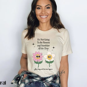 May include: A white t-shirt with a graphic of two cartoon flowers and the text "It's Not Going To Be Flowers and Sunshine All the Time. But they will be back again."