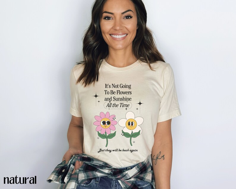 May include: A white t-shirt with a graphic of two cartoon flowers and the text "It's Not Going To Be Flowers and Sunshine All the Time. But they will be back again."