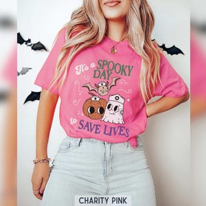 Puede incluir: Una camiseta rosa brillante con la frase "It's a Spooky Day to Save Lives" e ilustraciones de Halloween. El dise&ntilde;o incluye un murci&eacute;lago, una calabaza y un fantasma con gorro de enfermera.