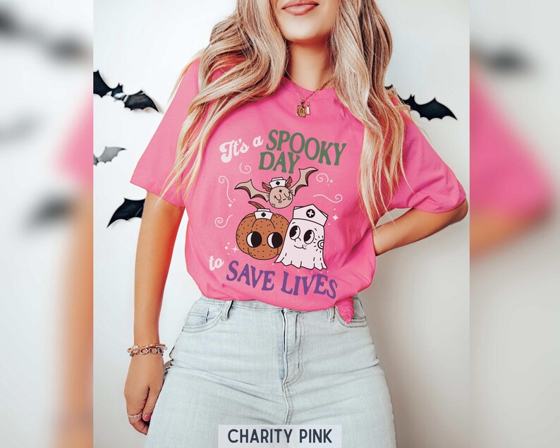 Puede incluir: Una camiseta rosa brillante con la frase "It's a Spooky Day to Save Lives" e ilustraciones de Halloween. El dise&ntilde;o incluye un murci&eacute;lago, una calabaza y un fantasma con gorro de enfermera.