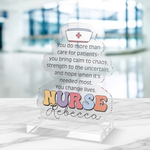 May include: A clear acrylic nurse-themed desk decoration. The design features a nurse's cap with a red cross, the word "NURSE" in colorful letters, and the name "Rebecca" in cursive. The text reads: "You do more than care for patients... You change lives."