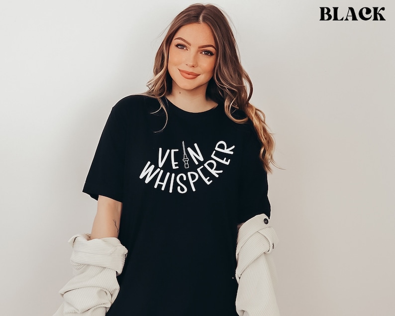以下が含まれることがあります： 白い文字で「Vein Whisperer」と書かれた黒いTシャツ。テキストは半円形で、中央に小さな針の画像があります。
