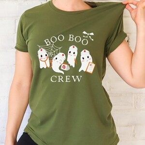 May include: Olive green t-shirt featuring a design of four ghosts dressed as nurses. The ghosts are wearing white hats with red crosses and holding medical supplies. The text "BOO BOO CREW" is printed below the ghosts.