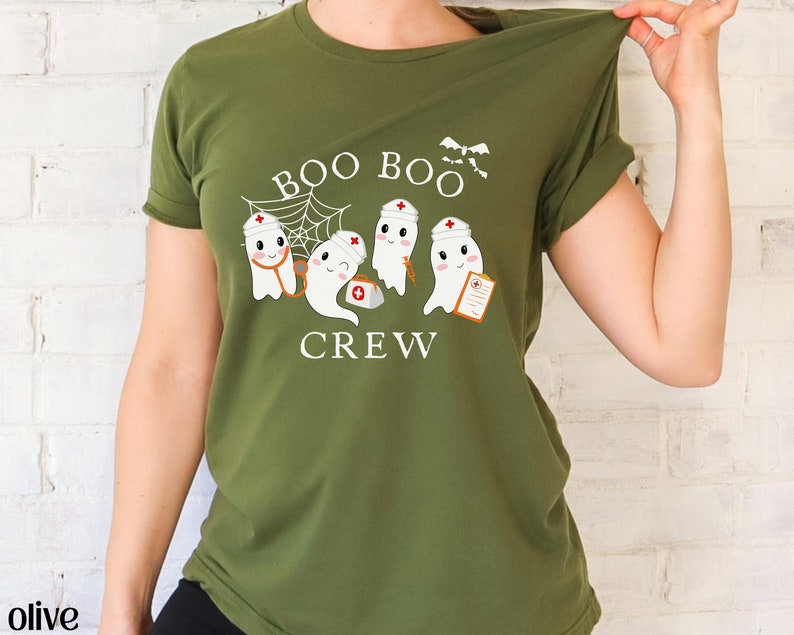 May include: Olive green t-shirt featuring a design of four ghosts dressed as nurses. The ghosts are wearing white hats with red crosses and holding medical supplies. The text "BOO BOO CREW" is printed below the ghosts.
