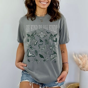 Op de afbeelding: Een grijze t-shirt met de tekst "BE KIND TO ALL KINDS" boven illustraties van insecten en vlinders in groentinten. De t-shirt heeft een relaxte pasvorm en korte mouwen.