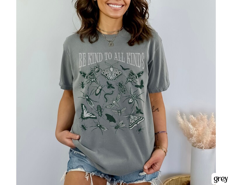 Op de afbeelding: Een grijze t-shirt met de tekst "BE KIND TO ALL KINDS" boven illustraties van insecten en vlinders in groentinten. De t-shirt heeft een relaxte pasvorm en korte mouwen.